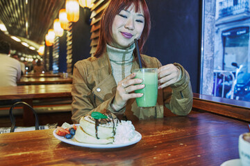 Portrait of gorgeous cheerful Chinese woman eating pancakes and drinking milkshake in cafe