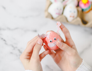 Children's hands hold one pink egg with glued eyes and stick stickers with bunny zigzag ears