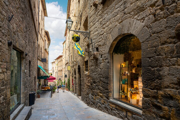 Tourists walk down a narrow cobblestone alley past shops and cafes in the historic medieval old town of the walled Tuscan city of Volterra, Italy.