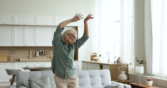 Pretty mature woman dancing feels happy, enjoy carefree leisure alone at home. Retired female moves to favourite music looks untroubled, spend free time in cozy living room. Pastime, hobby of retiree