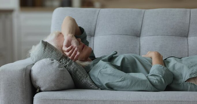 Close up weak or sick aged woman lying down on sofa at home looks exhausted or depressed, relieving fatigue, rest on couch in living room, having migraine, headache or sleep disorder, having day nap