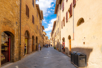 Tourists walk down a narrow, long cobblestone street of shops and cafes in the historic medieval old town of the walled Tuscan city of San Gimignano, Italy. - obrazy, fototapety, plakaty