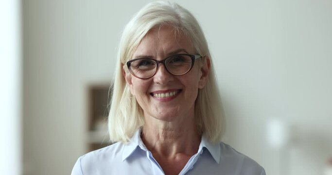 Happy mature businesswoman in casual blouse, head shot. Professional occupation person, businesslady portrait. Blond woman in glasses pose standing alone indoor smile looking at camera, close up face