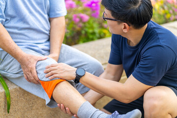 Elderly man suffering knee pain during jogging exercise with his son at park in the city. Adult man gives first aid to father with knee injury. Family relationship and older people health care concept