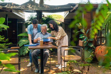 Asian couple surprised elderly father with Birthday gift at outdoor cafe restaurant on summer holiday vacation. Family relationship, celebrating father's day and older people health care concept.