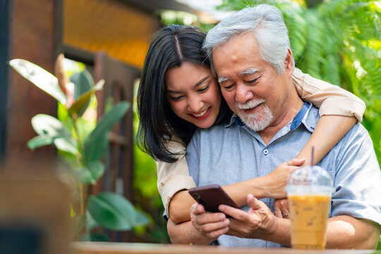Happy Asian woman teaching elderly father using mobile phone application during having lunch together at restaurant on summer vacation. Family relationship older people mental health care concept.