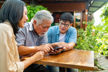Happy Asian couple teaching elderly father using mobile phone application during having lunch together at restaurant on summer vacation. Family relationship older people mental health care concept.