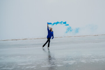 A caucasian woman is skating on a frozen lake holding colored blue smoke in her hands.