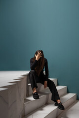 Chronic fatigue syndrome, concept. A young woman is sitting wearily on the steps, a failure when moving up the career ladder, professional burnout. Turquoise wall, copy the space from above.