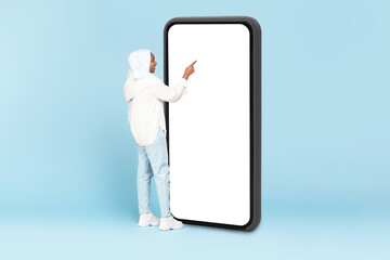 Muslim woman touching huge display panel of smartphone with blank white screen, standing on blue...