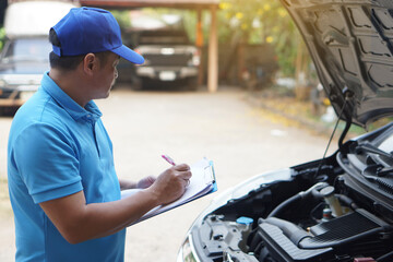 Naklejka premium Asian man mechanic wears blue cap and blue shirt, holds paper, checking and analyzing car engine under the hood. Concept, Outdoor car inspection service. Claim for accident insurance. 