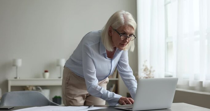 Attractive successful aged businesswoman in glasses typing on laptop smile look at camera, portrait of professional employee, teacher prepare work on computer, pose at workplace. Business, modern tech