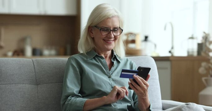 Middle-aged woman sit on sofa, hold smartphone, enter credit card details, makes purchases remotely, buys goods, pay for services use application. Happy female shopper use instant easy mobile payment