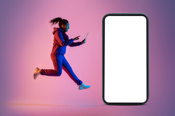 Young black lady jumping with digital tablet next to giant smartphone with blank white screen on pink neon background