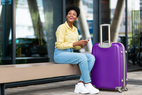 Black Female Traveler Using Cellphone Posing With Suitcase At Airport
