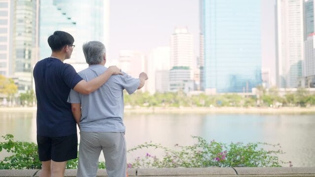 Asian adult son and elderly father looking urban cityscape during jogging exercise together at park. Retired man enjoy outdoor sport workout. Family relationship and older people health care concept.