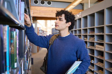 Young man, a university graduate student, selecting a book from a bookshelf in the library campus