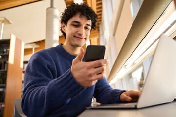 Hispanic young male student checking phone while studying online using laptop in the library campus