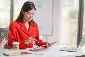 Female attractive asian banker accountant wearing red formal suit calculate the cash bill of audit...