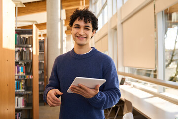 Handsome Hispanic male student with tablet, smiling, at camera, visiting a modern library campus