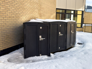 A row of adjoined rectangle shaped bicycle storage lockers against a brick wall with white snow on the ground.  The tough bike compartments have metal handles. 