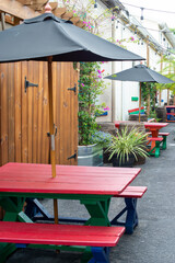 Fototapeta na wymiar The exterior patio garden of a restaurant, coffee shop or microbrewery style business with red wooden picnic tables, black umbrellas, and green potted plants. 