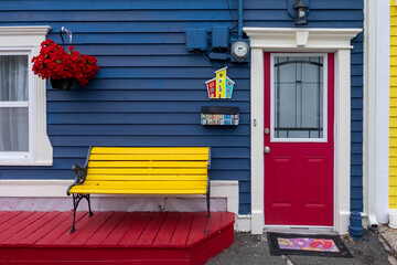 The entrance to a blue wooden house with a vibrant red door and glass window. There's a hanging red...