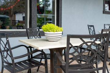 Tables and chairs on a patio terrace of a restaurant off a sidewalk. Each table has a flower pot with greenery. The cafe wall is grey with black trim. The garden furniture is black and white metal. 