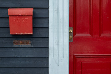 A bright red metal mailbox or letterbox on a deep blue wooden wall with a red door.  The mailbox has a curved shape cover. There's a shiny door handle, brass metal. The trim house is white in color.