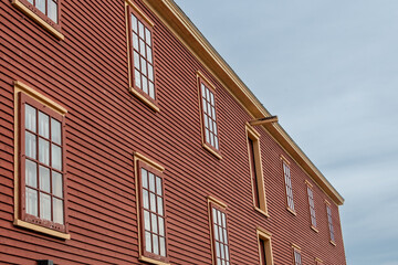 The exterior of a rusty red colored vintage warehouse with a wooden clapboard horizontal siding covered wall. There are two rows of windows with yellow trim, multi-glass panes, and a flat roof. 