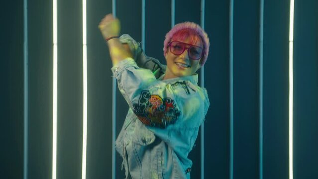 Trendy happy young woman vlogger listeng music, dances and recording videostream video for social media at home. Disco dancer in night club at light wall. Creating dancing video using tiktok app. 