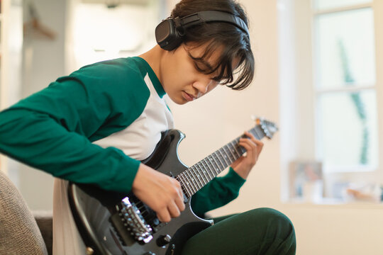Asian Teen Guy Playing Electric Guitar Wearing Headphones At Home - Powered by Adobe