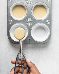 Hand scooping vanilla cake batter into a cupcake tin, An ice cream scoop being used to portion cake batter into a cupcake pan, process of making cupcakes