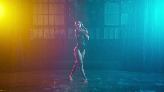 Hot dancer sitting on wet floor under rain garage background, moving sexually making erotic show teal orange spotlights. Seductive woman showing sexy body performing striptease cyberpunk urban stage