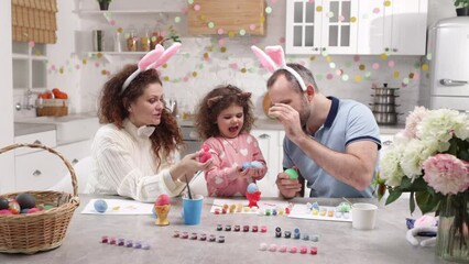 Portrait of caucasian family in bunny ears celebrating traditional Easter. Father and mother showing their cute little toddler how to eat traditional colorful eggs. High quality 4k footage