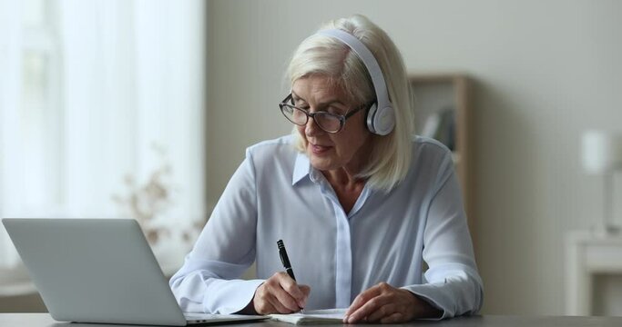 Attentive mature woman in headphones e-learn, improve foreign language knowledge take part in on-line class with tutor, make notes, write exercise, studying use modern technology. Education, tuition