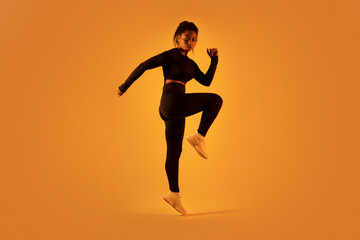 Fototapeta na wymiar Sporty black woman jumping and doing stretching exercise over orange neon light background, full length