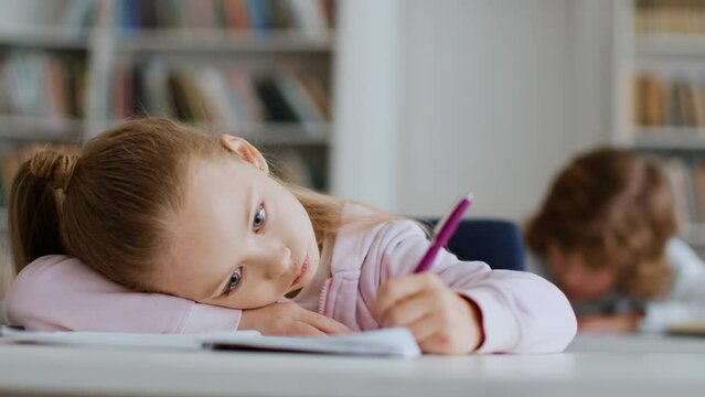 Boring education. Little schoolgirl lying on table and writing task in copybook, uninterested to learning, free space