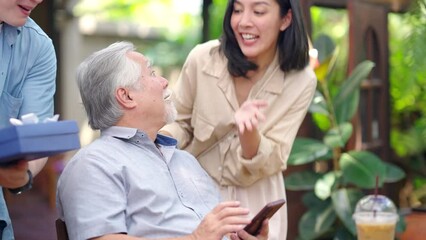 Asian couple surprised elderly father with Birthday gift at outdoor cafe restaurant on summer holiday vacation. Family relationship, celebrating father's day and older people health care concept.