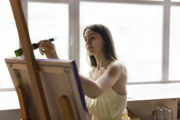 Engaged artist girl painting picture in art school, drawing on canvas, using paintbrush, enjoying creative work, hobby, inspiration, training artistic skills, standing against large window