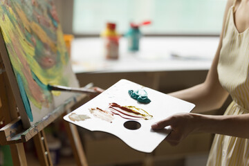 Artist using craft palette for drawing picture, holding pad with colorful acrylic paints, stroking on canvas in background, working at easel. Close up shot of female hands, appliance