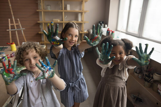 Cheerful cute multiethnic kids posing at camera in creative craft studio, showing painted palms, smiling, laughing, having fun. Pupils enjoying leisure time on artistic class