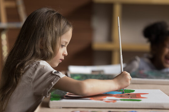 Focused cute pretty art school pupil girl drawing cartoon animal on canvas, studying creativity on painting class, holding paintbrush, sitting at big table in classroom. Side view