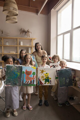 Multiethnic group of cheerful children and art school teacher holding painted pictures, showing artworks, looking at camera, smiling, laughing. Full length vertical shot