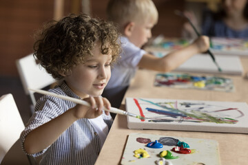 Pretty pupil kid boy painting on class, studying art in artistic school for children, drawing abstract picture on canvas at table, using paintbrush, palette with oil, acrylic paints