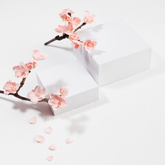 Spring stage mockup - two square podiums for presentation cosmetic products, goods, twig of tender pink sakura flowers, petals on white wood background in japanese traditional style, top view.