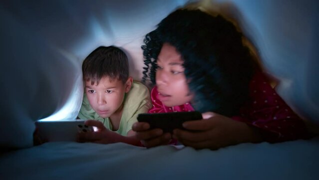 Curious preschool kid boy using digital smartphone technology device in bed under blanket at night. Small child and mother competition in mobile online game on mobile phone. Children tech addiction 4K