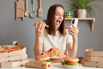Extremely happy overjoyed Caucasian woman with brown hair wearing white t shirt sitting in kitchen at table and holding slice of pizza in hands, having dinner with junk food, using mobile phone.