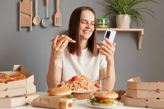 Image of smiling happy woman with brown hair wearing white t shirt sitting in kitchen at table, eating pizza and using cell phone, checking social networks while having snack.