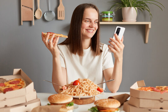 Photo of cheerful Caucasian young adult woman with brown hair wearing white t shirt sitting in kitchen at table and looking at smart phone display, having delicious snack, eating pizza.